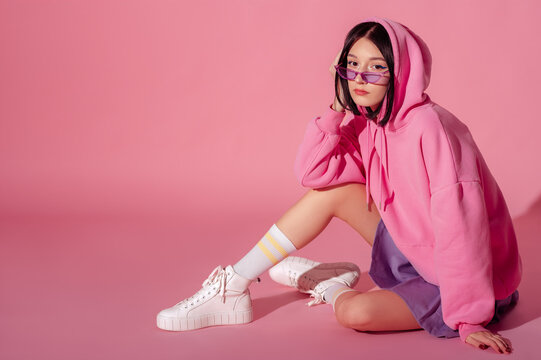 Fashionable confident woman wearing trendy pink hoodie, purple  sunglasses, mini skirt, white high top sneakers, striped socks, sitting, posing on pink background. Copy, empty space for text