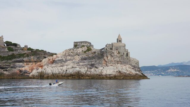 Sailing by famous historic church Chiesa di San Pietro and Doria Castle in Porto Venere in Liguria, Italy. A small motor boat sails past a rock.