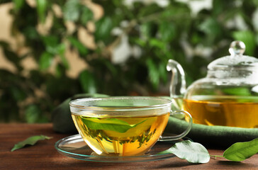 Fresh green tea in glass cup with saucer, leaves and teapot on wooden table