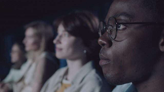 Selective focus close-up of young Black man wearing eyeglasses watching movie adn eating popcorn in cinema, his Caucasian girlfriend whispering something to him