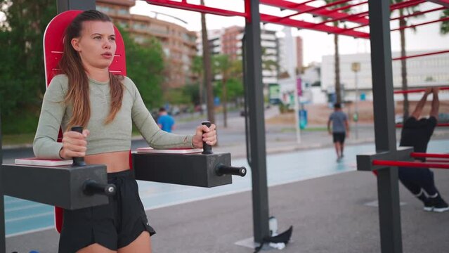 Fitness, sport, sportive people concept - young woman doing abdominal exercise on parallel bars in outdoor gym. Caucasian fitness female training abs outside on sports ground. 