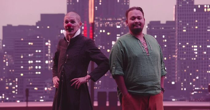 Portrait of the multiethnic team, a pacific islander man with his best friend clown standing and looking at the camera with city lights at night in the background.