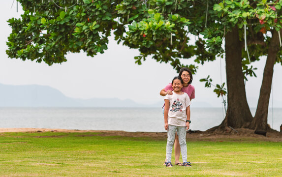 Portrait Asian mother and little daughter stand on grass next to the beach, happy face with smile, wide image background of big tree, sea and mountain, space for text.