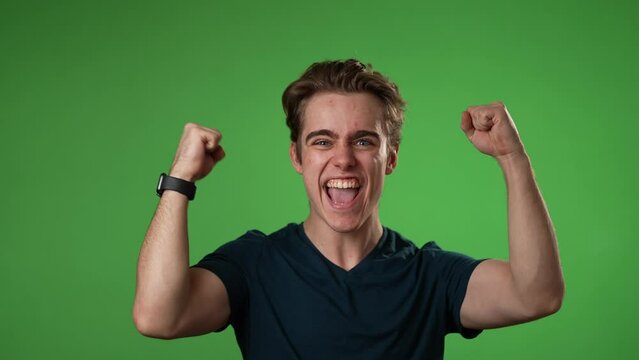 Excited jubilant overjoyed young man 20s doing winner gesture celebrate clenching fists say yes isolated on green screen background studio portrait in slow motion