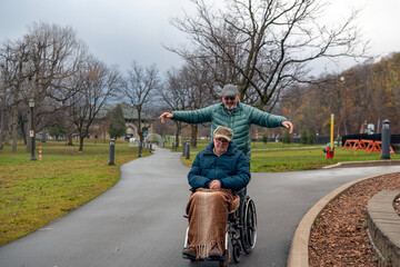 Naklejka premium A senior gay man in a wheelchair on an autumn day. His husband who should be pushing the wheelchair has his arms in the air and letting the chair go down the incline path.