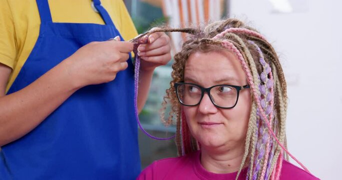 Unprofessional hairdresser cuts a strand of tangled curly hair with scissors, while unwinding the box braid of a young blonde woman with glasses. Use of substandard services.