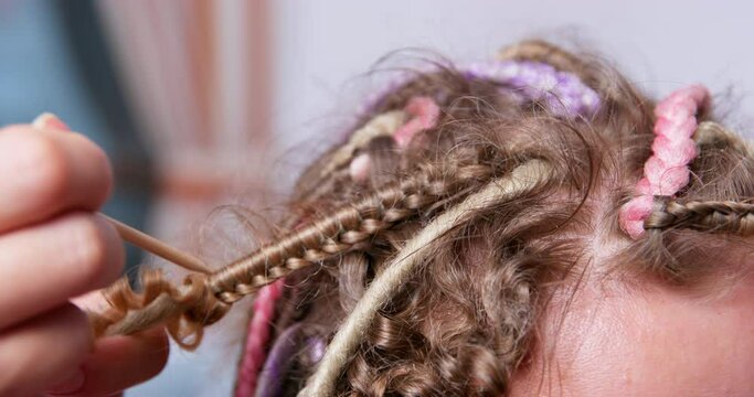 Hairdresser-stylist in a blue apron unwinds multicolored box braids to client using special wooden tool, after which the hair becomes curly, side view, close up. Master at work in a beauty salon.
