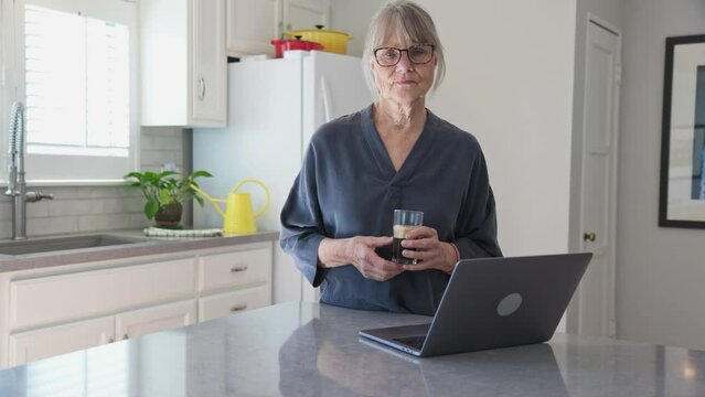 Dolly shot of Senior Caucasian woman using laptop computer in kitchen while drinking iced coffee. Medium shot of older white lady working on pc at the counter. Slow motion 4k