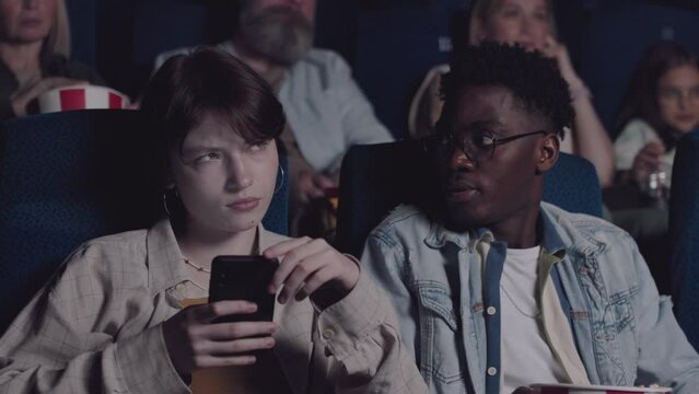 Young gen Z woman texting something on smartphone or surfing internet while watching movie in cinema with her boyfriend