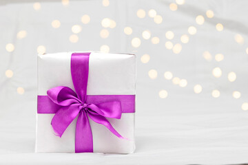 Decorative white gift box with a large purple bow against a background bokeh of lights