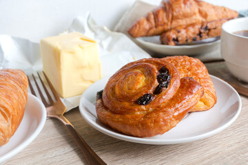 Cinnamon rolls, buns on white dish with raisins, croissant and butter on gray wooden table. Variety of homemade buns pastry. close up of cinnamon rolls.