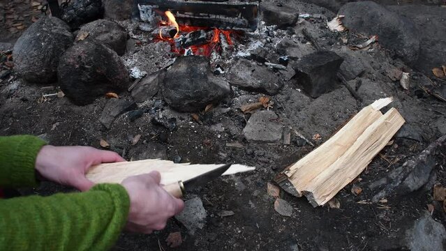 White male whittle chips from wood log with finnish puukko knife. Green shirt. Camp fire on the background. Static footage.