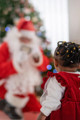 back view of the little African girl looking at Santa Claus in her living room at home, selective focus