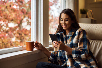 Happy woman using smart phone while drinking coffee by window at home.