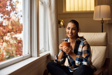 Smiling woman enjoys in cup of tea while relaxing in armchair by window.