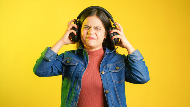 Beautiful young woman listening to music using headphones, Asian Indian girl removing her earphones to avoid loud music, ear pain concept, isolated over colour background studio shot