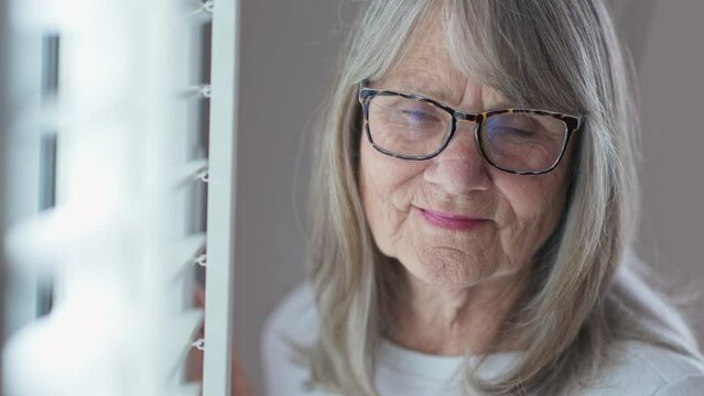 Close up portrait of friendly looking senior Caucasian woman in glasses looking at camera. Retired older white woman by a window. Slow motion 4k