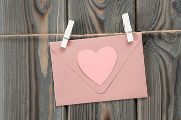 Pink cute paper envelope with heart shape in the center hanging on rope with clothespins on dark wooden background. Love letter on holidays. Handmade postcard for valentines day. Copy space, mockup