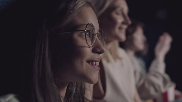 Selective focus close-up of cheerful preteen Caucasian girl wearing eyeglasses eating enjoying watching movie in cinema
