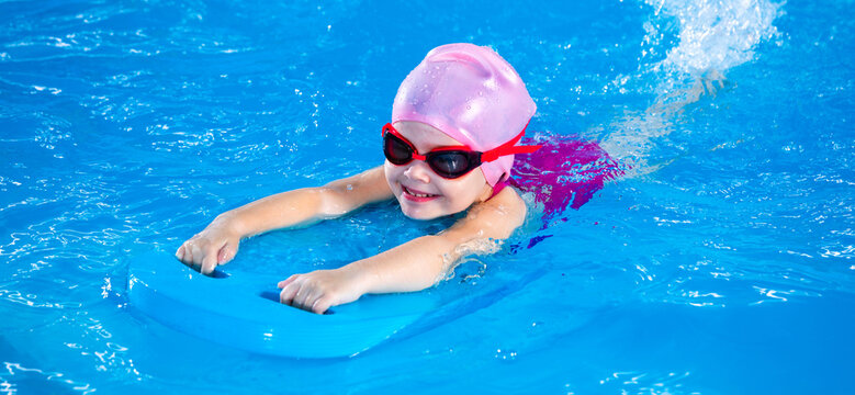 Panorama of Smiling little girl learning to swim in pool with flutterboard during swimming class