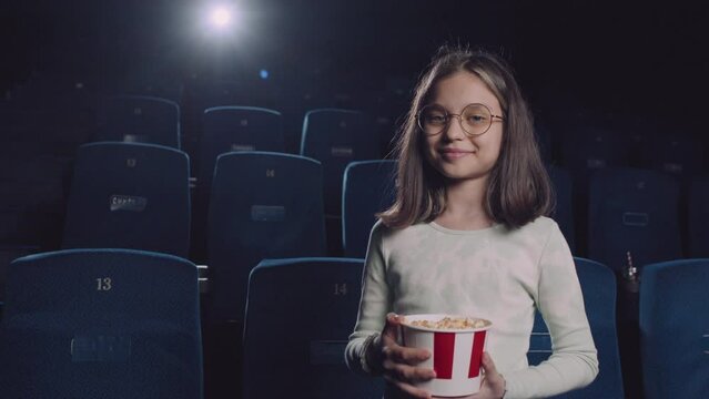 Medium length portrait of preteen Caucasian girl wearing eyeglasses holding bucket of popcorn standing in cinema hall against navy blue seats