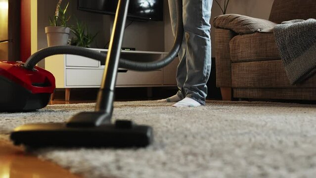 Home cleaning service and chores. Household woman vacuums carpet near sofa in apartment. Low angle view of vacuum cleaner and female legs. Tracking left shot.