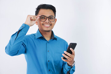 smiling asian employee man holding phone and holding the edge of the glasses wearing blue shirt isolated on white background