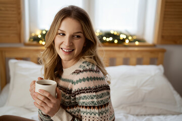 Caucasian young woman sitting on bed with cup of coffee and turning
