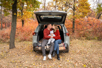 Parents in red sweaters sit in the trunk of a car and hug their baby at an autumn photo shoot