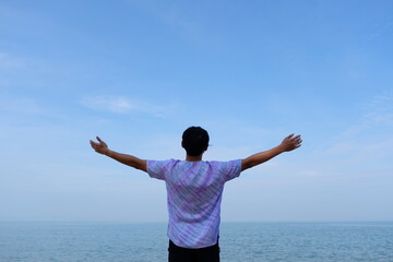 man on beach hold hands arms up, rear view guy wearing summer hat, standing back looking to sea blue sky horizon, vacation concept of freedom travel ocean