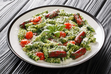 Hearty lunch Fusilli pasta with sausages, tomatoes, green pesto sauce and cheese close-up in a plate on the table. Horizontal
