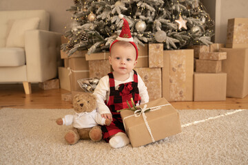 A cute baby in a Santa hat sits and smiles under a festive Christmas tree with a gift and a teddy bear