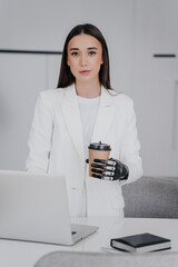 Vertical shot of beautiful brunette girl in white suit with bionic hand prosthesis holds cup of...