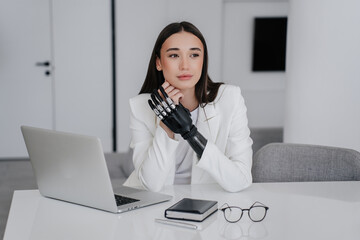 Beautiful brunette caucasian woman in white suit with artificial arm prosthesis sitting at desk...