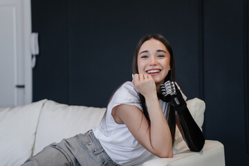 Fototapeta na wymiar Smiley hispanic young pretty woman leaning on bionic artificial hand, sits on sofa looks at camera confidently. Satisfied beautiful girl in love relaxing at home. Rehabilitation after injury.