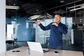 Naklejka na ściany i meble Businessman celebrating achievement, asian man dancing and singing at workplace, man in shirt working inside modern office using laptop at work.