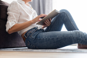 Woman in white blouse and jeans sitting on a floor at home and using a tablet.