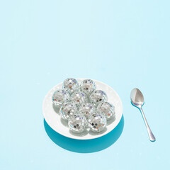 Disco balls in white plate and silver spoon on bright blue background. Minimal retro food party concept. Surreal festive artistic idea. New Year celebration aesthetic.