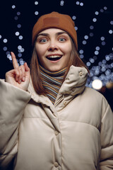 Surprised woman in warm winter clothes pointing up and expressing astonishment against garlands in city at night 