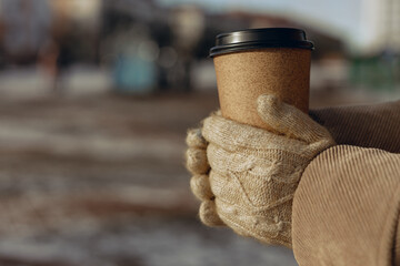 Fototapeta na wymiar Crop woman in knitted gloves holding paper cup of takeaway coffee and warming hands on sunny day in winter in city