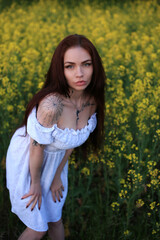Naklejka na ściany i meble a beautiful fashionable girl with red hair stands in a rapeseed field in a white dress, with tattoos on her body against a background of yellow flowers