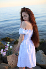 Fototapeta na wymiar a beautiful young fashionable girl with red hair in a white dress stands in summer by the water of the sea ocean with tattoos on her body and a bouquet of flowers