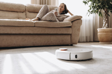 Woman is sitting on couch and operating white robot vacuum cleaner via wi-fi. Girl wearing home clothes resting relaxing lying on sofa with smartphone. Smart home - obrazy, fototapety, plakaty