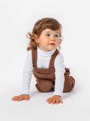 Caucasian toddler 2 years old with long curly hair on white background in a brown jumpsuit and white turtleneck crawls on his knees on the floor on white background. The head is turned to the side.