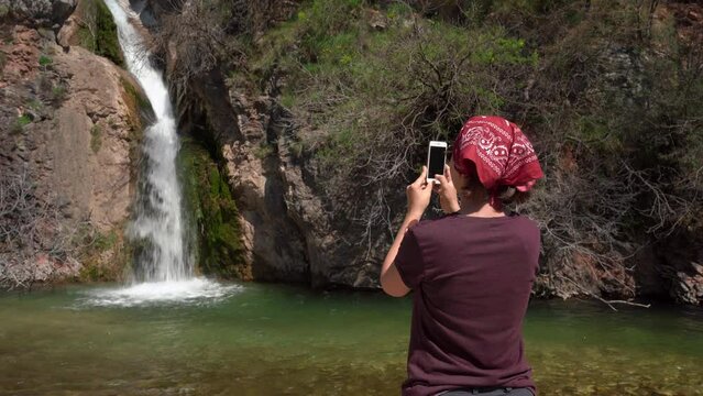 Tourist in red bandana takes photos of waterfall during camping stop, using mobile phone, view from the back. Young female blogger makes content for her travel vlog.