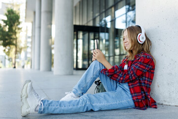 Young smiling beautiful hipster happy teen girl in city street,speaking on smartphone,headphones, jeans, backpack, positive, spring trend, urban teenage style
