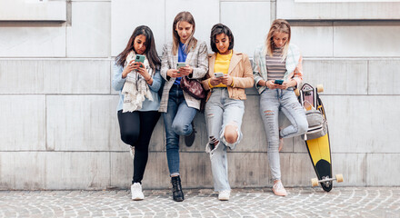 generation z multiethnic girls and friends using cellphone leaning against wall in university - obrazy, fototapety, plakaty