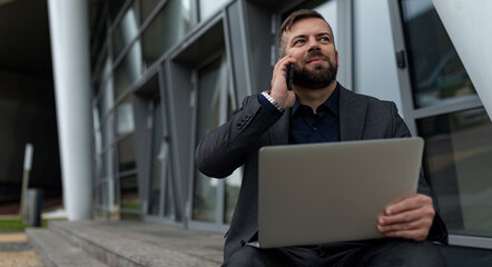 businessman talking on a mobile phone with a laptop on his knees while sitting at the entrance to the office building