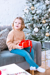 Naklejka premium cute happy little girl with curls is sitting on chair near Christmas tree with gift box in her hands, New Year and Christmas family concept