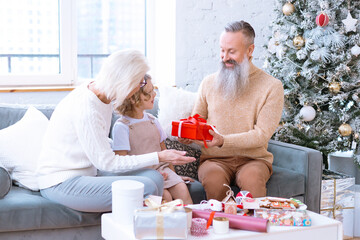 Christmas family grandmother, grandfather and granddaughter sit on sofa near Christmas tree and preparing handmade New Year presents, pretty girl and active happy seniors give each other gift box